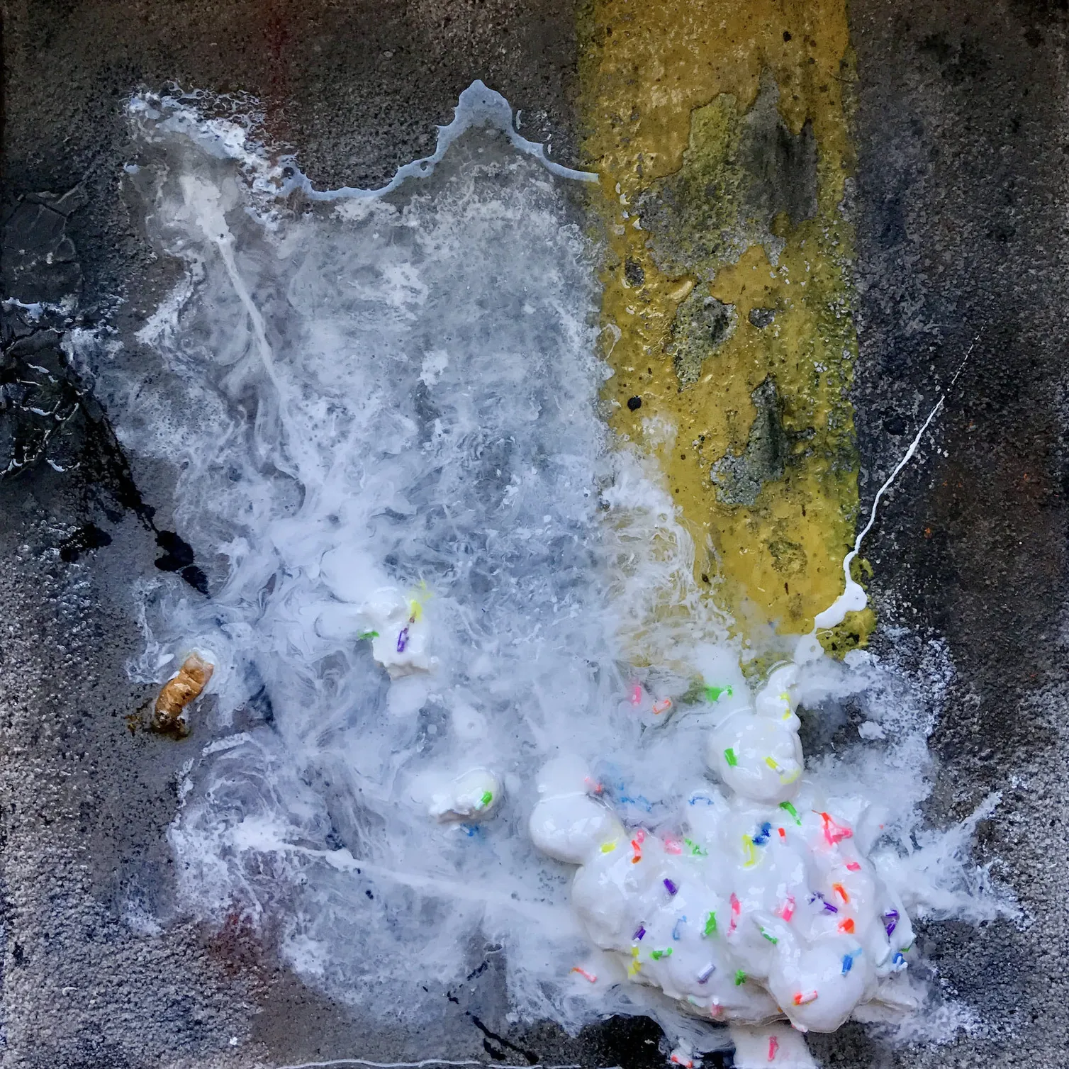 A three-dimentional painting of a ball of white ice cream, with rainbow sprinkles, splattered across dark, textural, parking lot asphalt. The splattered dessert overlaps an amber yellow line denoting a parking stall towards the top of frame. There is a discarded, smashed cigarette butt laying in a puddle of splattered ice cream to the left of the ice cream ball.